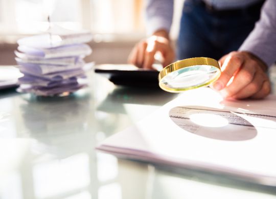 Businessperson's Hand Analyzing Bill Through Magnifying Glass